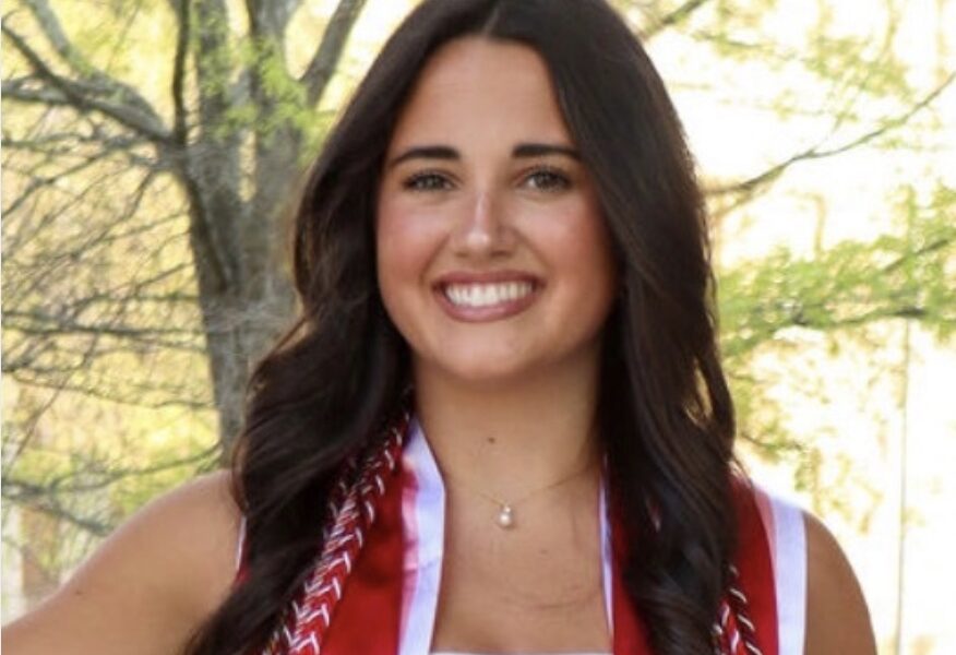 Young woman in a white dress wearing a red graduation stole, smiling outdoors.