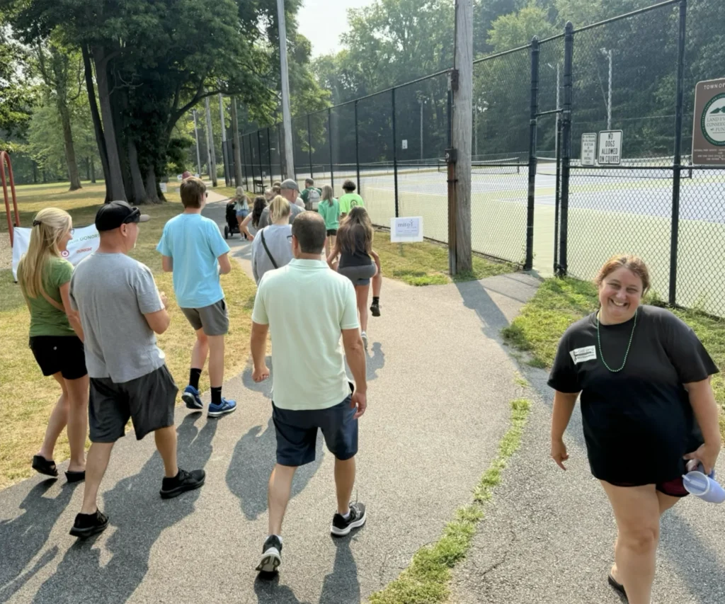 2025 Rochester Energy Walk 1 MitoAction - MitoAction Group of people walking along a park path beside a chain-link fence tennis court, with trees overhead and a smiling woman in the foreground right. MitoAction