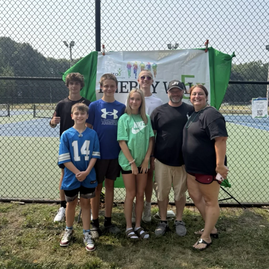 2025 Rochester Energy Walk 2 MitoAction - MitoAction Group of people posing in front of a banner on a tennis court, behind a chain-link fence. MitoAction