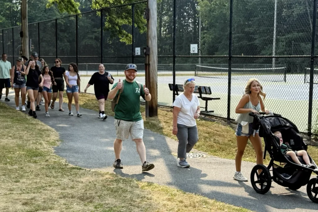 2025 Rochester Energy Walk 3 MitoAction - MitoAction Group of people walking along a park path beside a chain-link fence tennis court; man in green shirt at front gives thumbs up. MitoAction