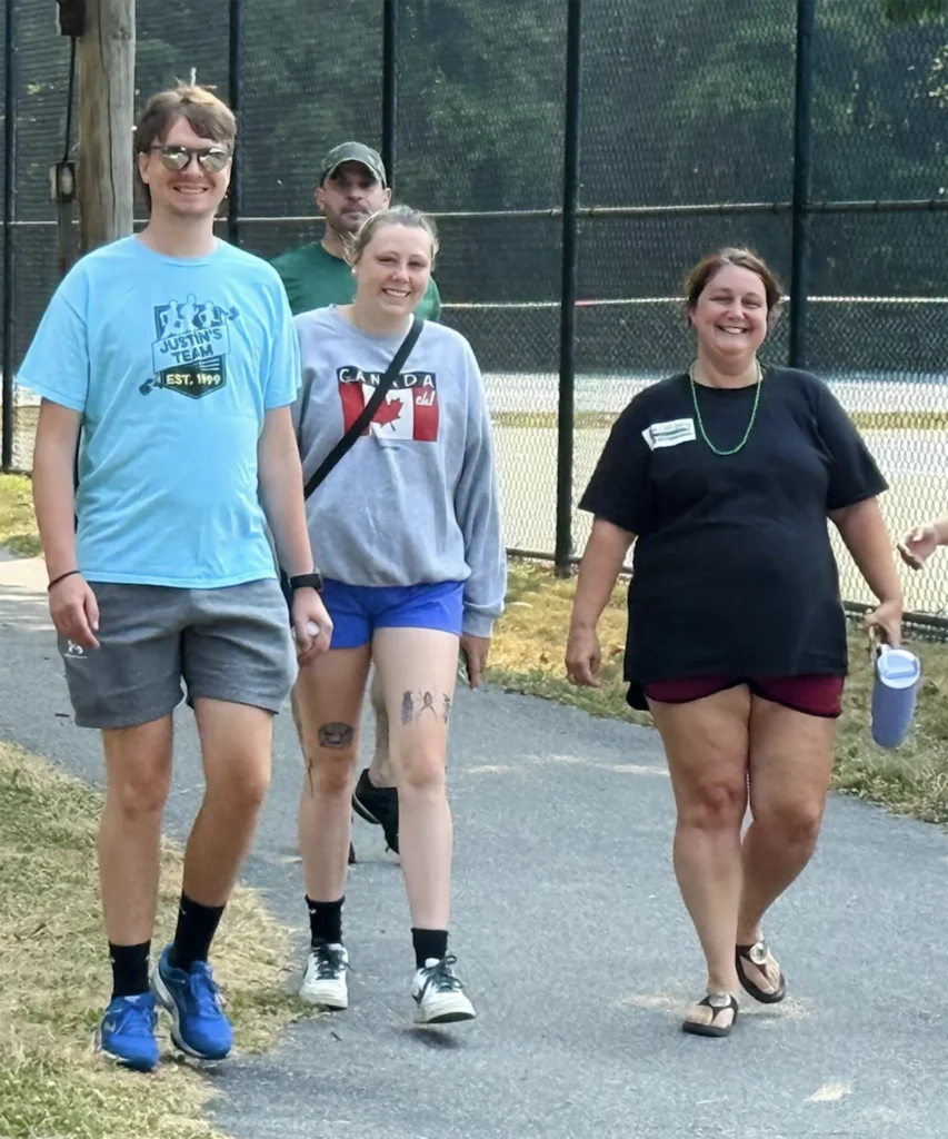 2025 Rochester Energy Walk 5 MitoAction - MitoAction Group of four adults walking side by side on a paved path beside a tall black fence, smiling at the camera. MitoAction