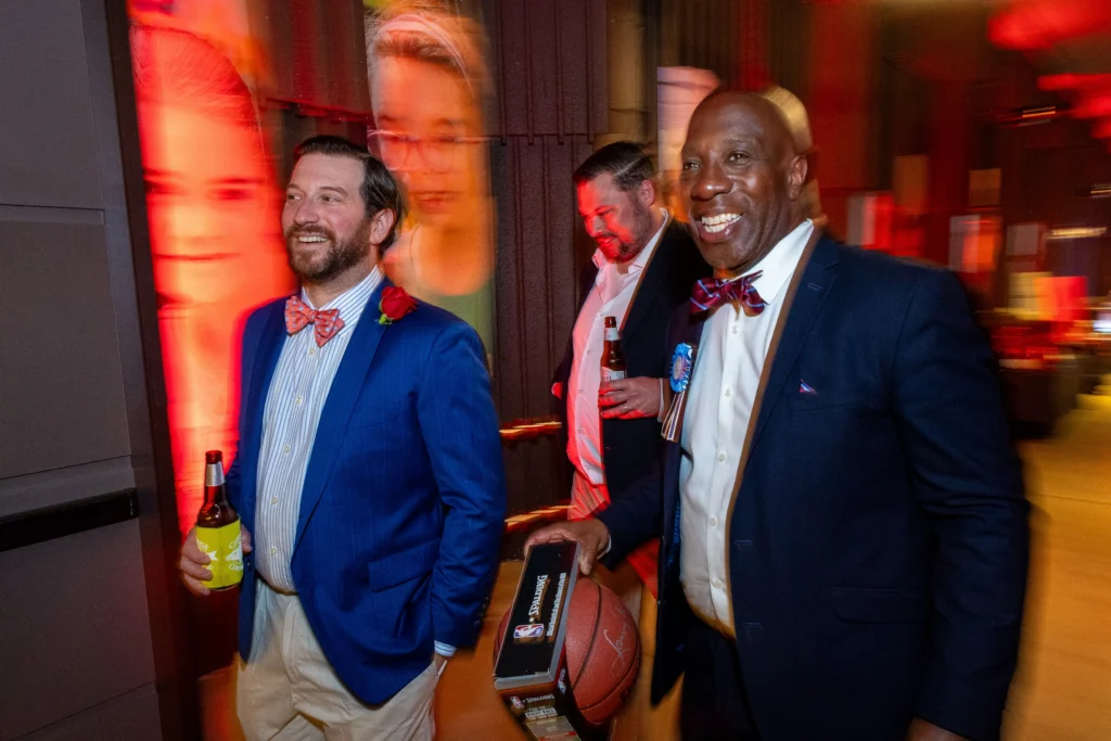 Three men in suits at a party; the man in front left wears a blue blazer and red bow tie, holding a beer, as others mingle under red lighting.  MitoAction
