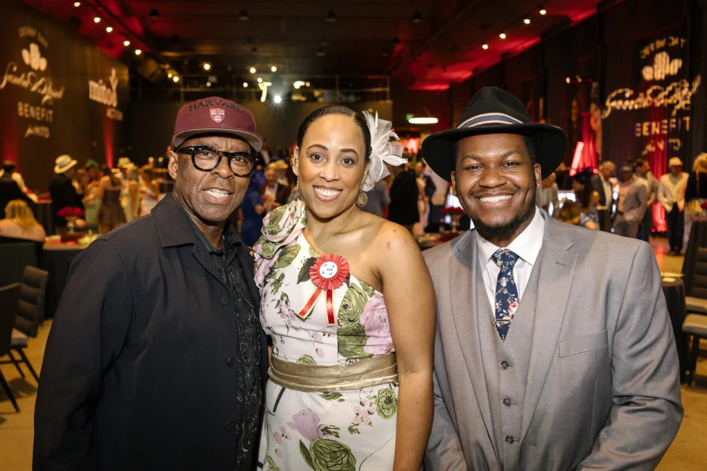 Three adults smile for a photo at a gala: man in a Harvard cap on the left, woman in a floral dress with a white flower in her hair in the center, and man in a gray suit and hat on the right, with a busy event hall in the background.  MitoAction