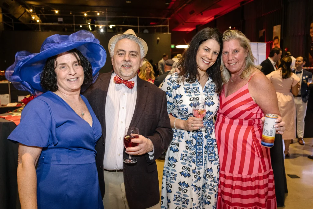 Four friends at a party pose for a photo, holding drinks and smiling at the camera.  MitoAction