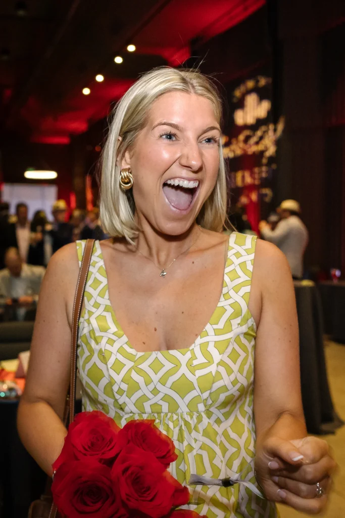 Woman smiling with mouth open, holding a bouquet of red roses at a lively indoor event.  MitoAction