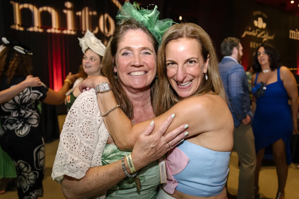Two women hug and smile at a lively party, wearing pastel dresses and festive headpieces.  MitoAction
