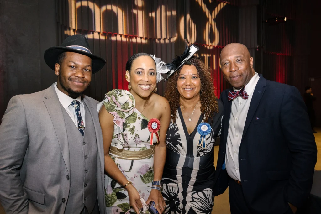 Group of four adults in formal attire posing for a photo at an event; two women wear floral/black-and-white dresses with decorative hats and ribbons, two men wear suits with ties.  MitoAction