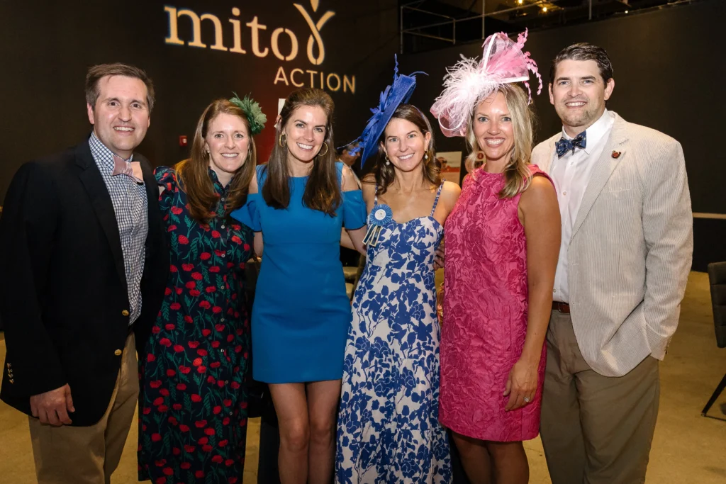 Group of six adults smiling for a photo at a formal event, wearing colorful dresses and hats. In the background, a sign reads   MitoAction'mito9 ACTION'.