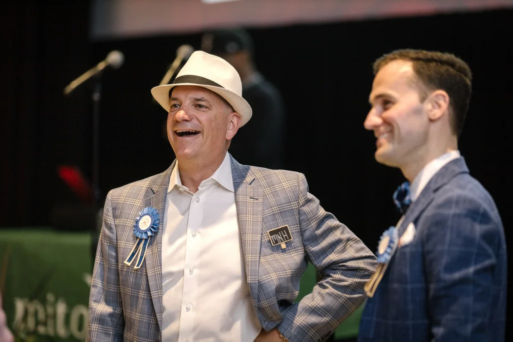 Two men in plaid blazers smiling at an event; the man on the left wears a white fedora and a blue rosette badge.  MitoAction