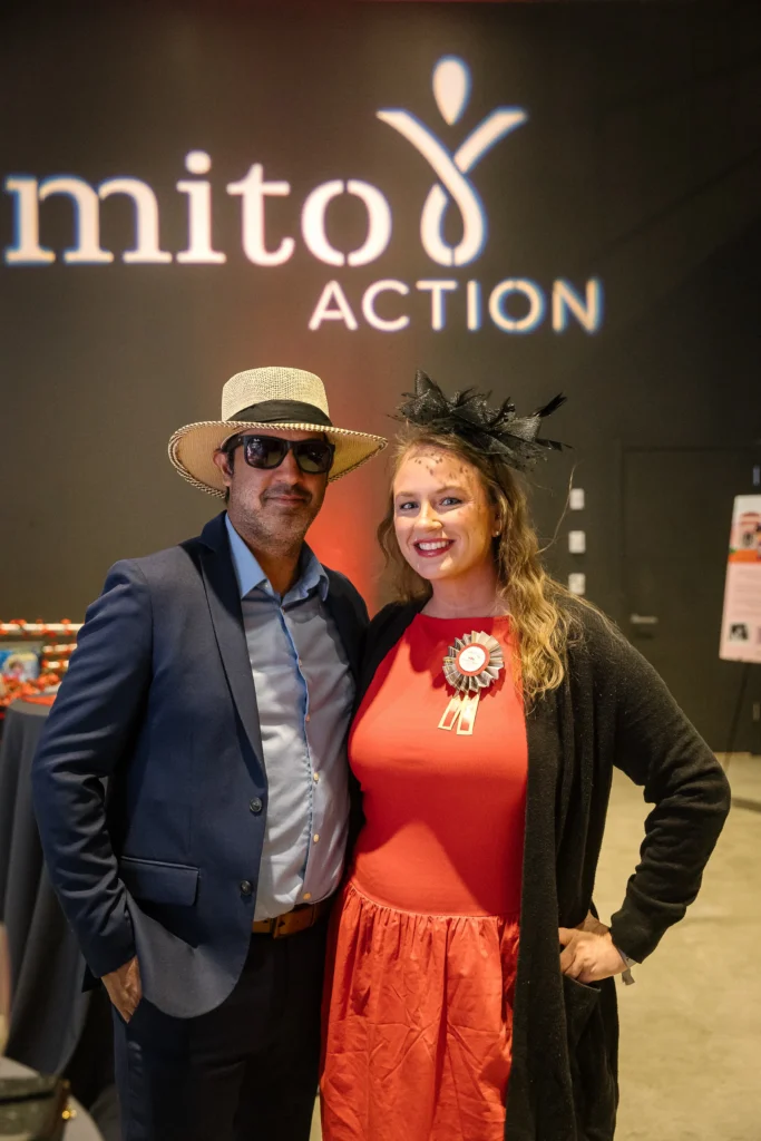 Two smiling adults pose for a photo at an event; man in a blue suit and straw hat with sunglasses, woman in a red dress with a black fascinator, backdrop reads   MitoAction'mito ACTION'.