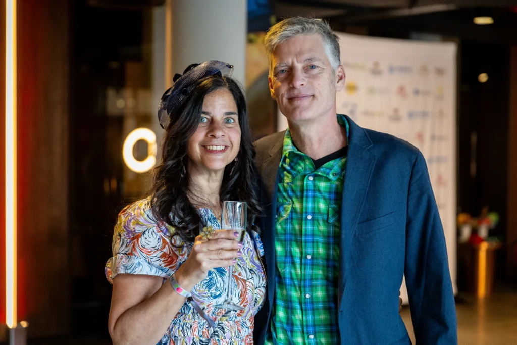 A smiling couple at a party, woman in a floral dress holding a champagne flute, man in a green plaid shirt and blazer beside her.  MitoAction