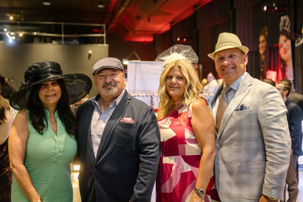 Four adults posing at an indoor party, wearing fancy hats and bright outfits, smiling for the camera.  MitoAction