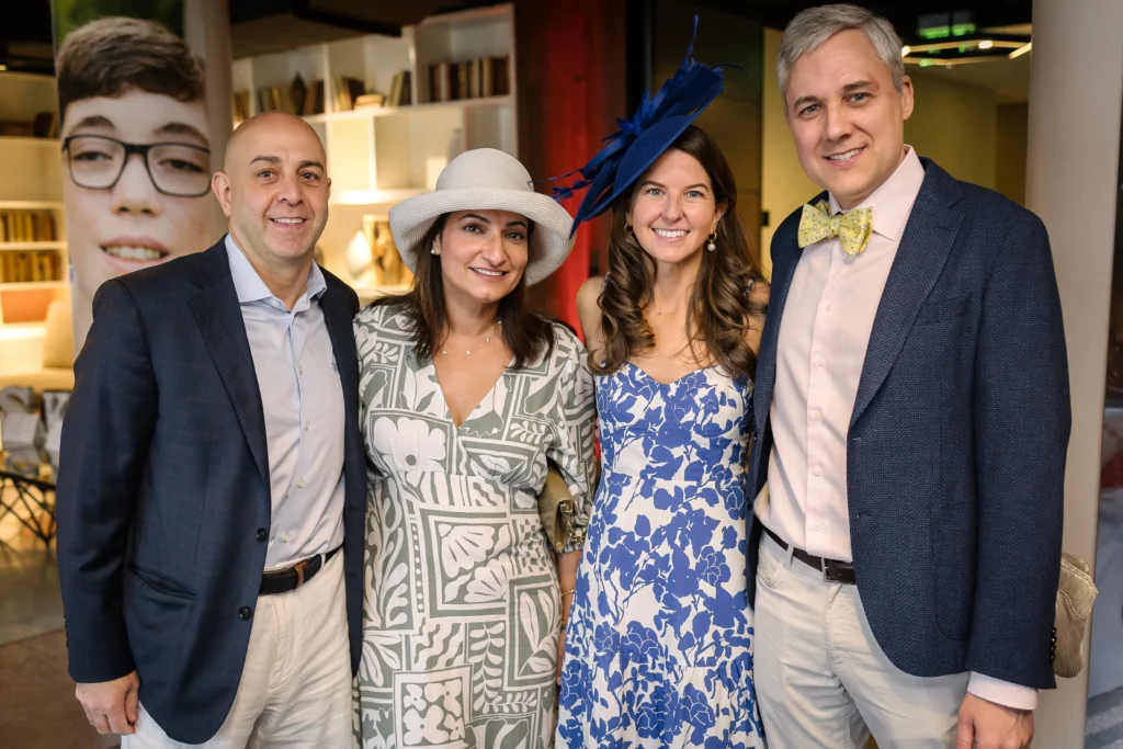 Four adults posing for a group photo at an indoor event; two men in blazers and two women in patterned dresses wearing hats, smiling at the camera with a bookstore-like backdrop.  MitoAction