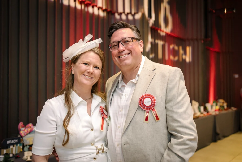 Two adults pose for a photo at an indoor event, both wearing ribbons and smiling. The woman wears a white blouse with a feathered hat; the man wears a light suit.  MitoAction