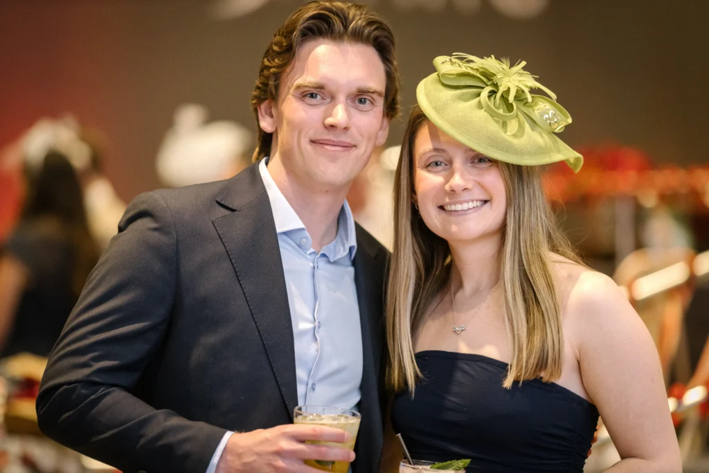 Smiling couple at a formal event; man in a suit holding a cocktail, woman in a navy dress with a lime-green fascinator hat.  MitoAction