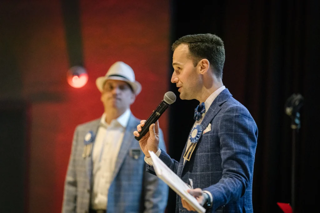 Man in a blue checkered suit speaks into a handheld microphone on stage, with another guest in a light suit and hat blurred in the background.  MitoAction