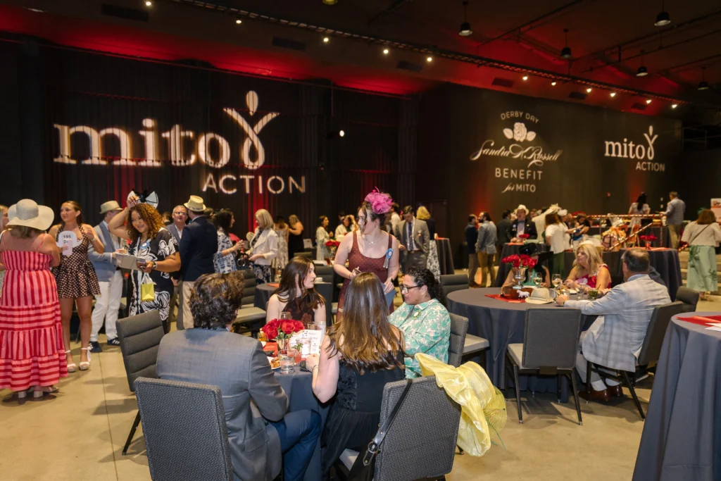 Busy fundraising gala with round tables, guests in formal wear, red roses centerpieces, and large banners reading ‘Mito Action’ in the background.  MitoAction