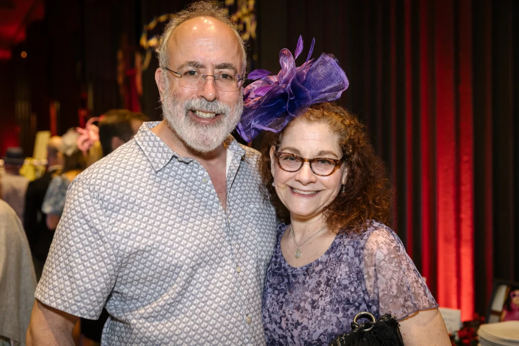 Smiling couple posing at an indoor event; man in patterned shirt, woman with purple fascinator and glasses, red curtains in background  MitoAction