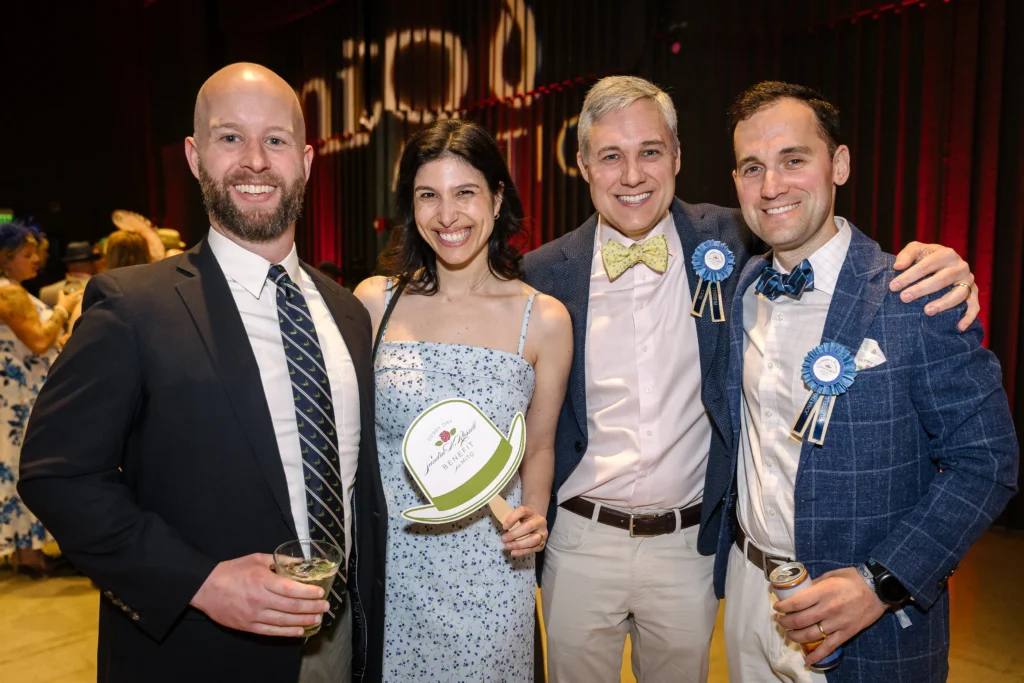 Four adults pose together at a social event, smiling, with drinks and ribbons on their outfits in a banquet setting  MitoAction