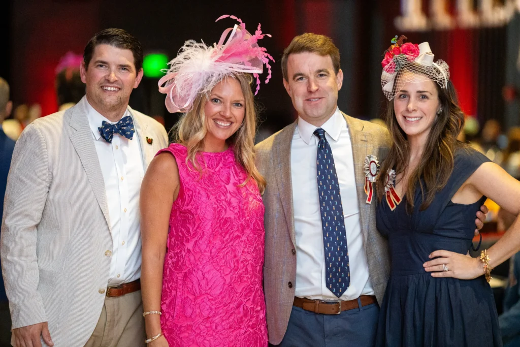 Four adults in formal wear posing for a group photo at a event, two men in suits and two women in bright dresses with decorative hats, smiling at the camera.  MitoAction