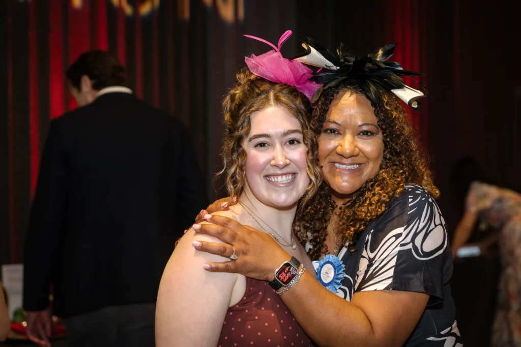 Two women smile and hug at a celebration; one wears a pink feather headpiece and the other a patterned dress with a blue ribbon badge.  MitoAction