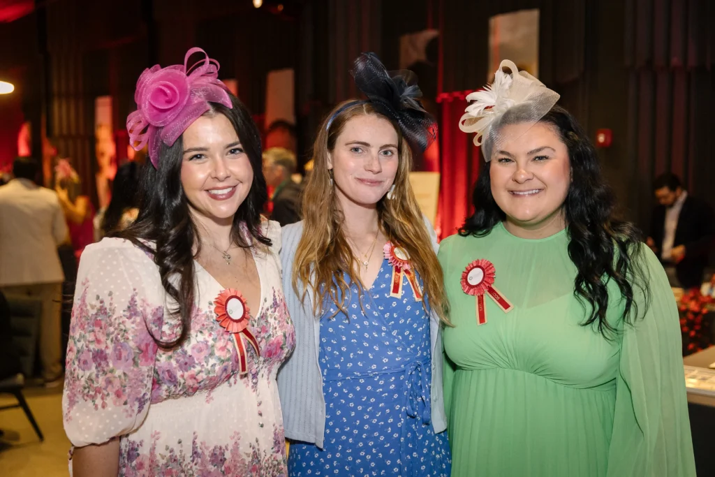 Three smiling women in floral and solid dresses wear decorative hats with ribbons at an indoor event, posing together.  MitoAction