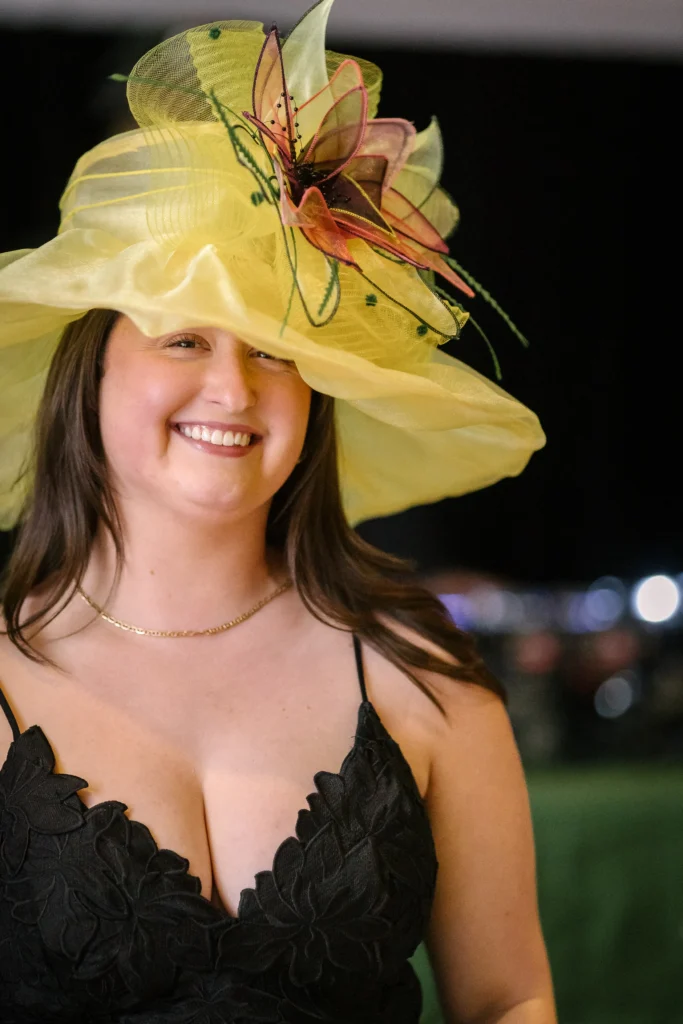 Smiling woman wearing a bright yellow fascinator with a decorative pink-orange flower, outdoors in a garden setting, wearing a black dress.  MitoAction