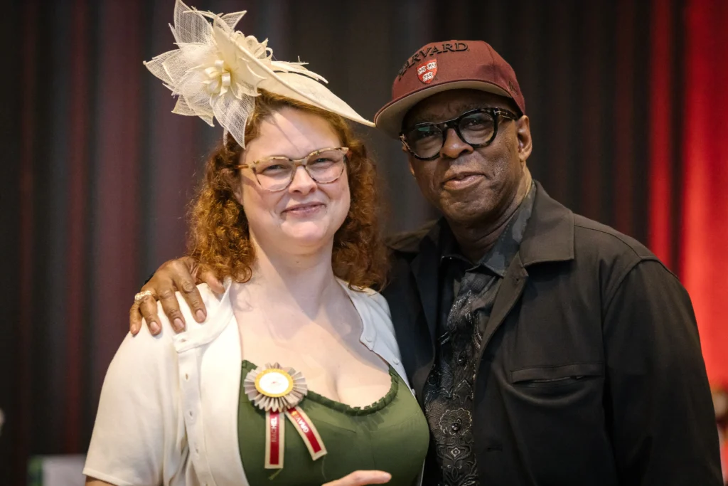 Smiling woman in green dress with a decorative cream hat and badge, standing beside a man in a maroon cap; they pose together on stage.  MitoAction