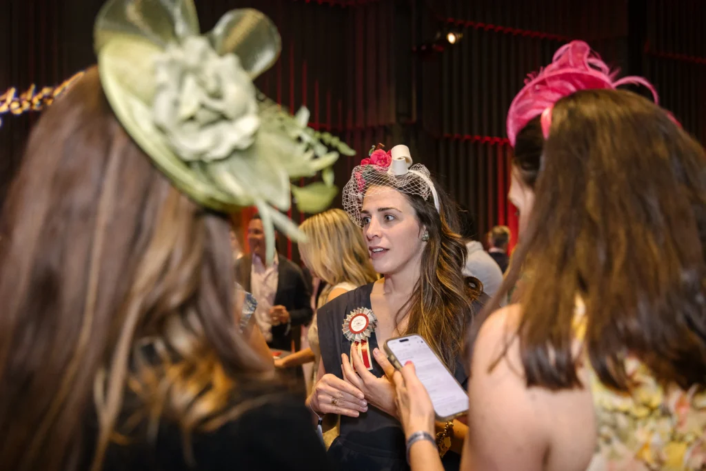 Center woman wearing a fascinator with a pink flower and birdcage veil talks to a crowd at a social event; she holds papers and wears a badge.  MitoAction