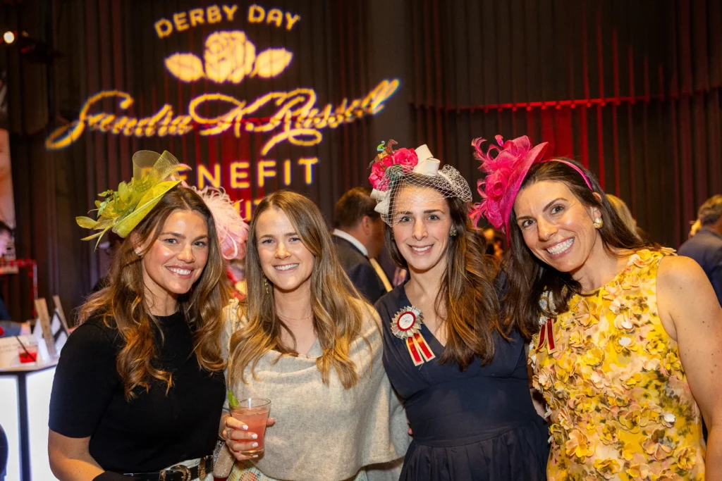 Four women in party outfits wearing elaborate hats smile for a group photo at a Derby Day event, indoors with a logo backdrop behind them.  MitoAction