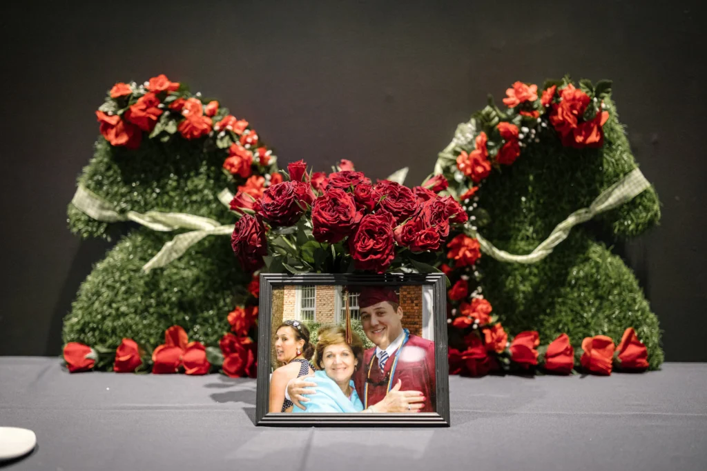 Framed group photo resting on a stage, surrounded by large red rose arrangements and greenery.  MitoAction