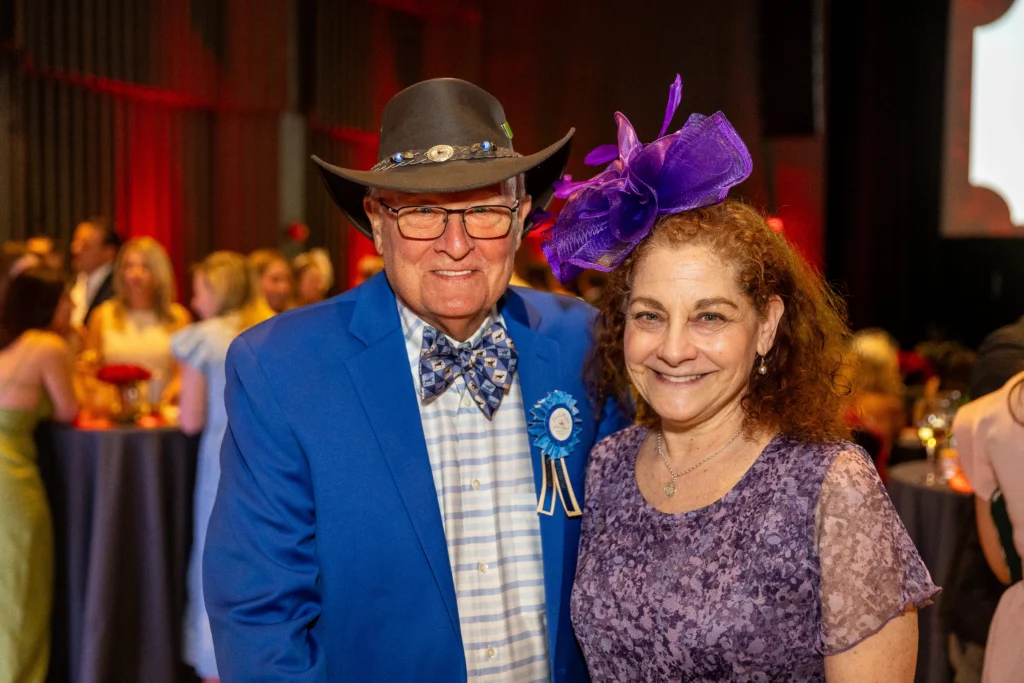 Smiling couple at a formal event: man in a bright blue suit and hat beside a woman wearing a purple fascinator, with guests in the background.  MitoAction