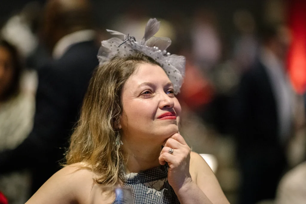 Woman wearing a lace and feather headpiece smiles, resting her chin on her hand at a dim indoor event.  MitoAction
