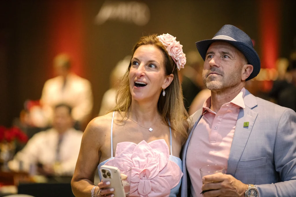 A smiling couple at a formal event, the woman wearing a pink dress and flower headband, holding a phone; the man in a light suit and fedora stands beside her.  MitoAction