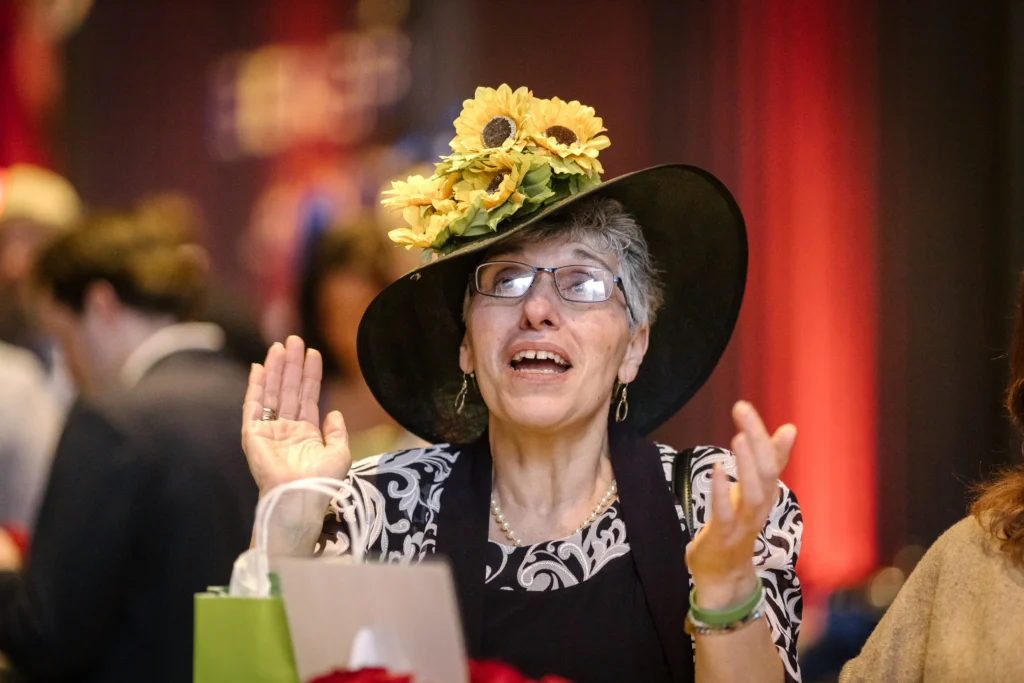 Woman in a black hat with bright sunflower decorations, smiling and clapping at a lively event.  MitoAction