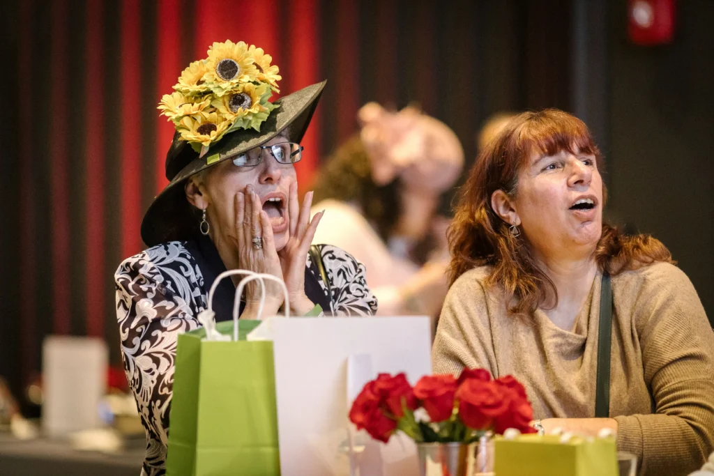 Two women seated at a table during a celebration; one wears a hat adorned with yellow sunflowers and looks surprised, with gift bags nearby.  MitoAction