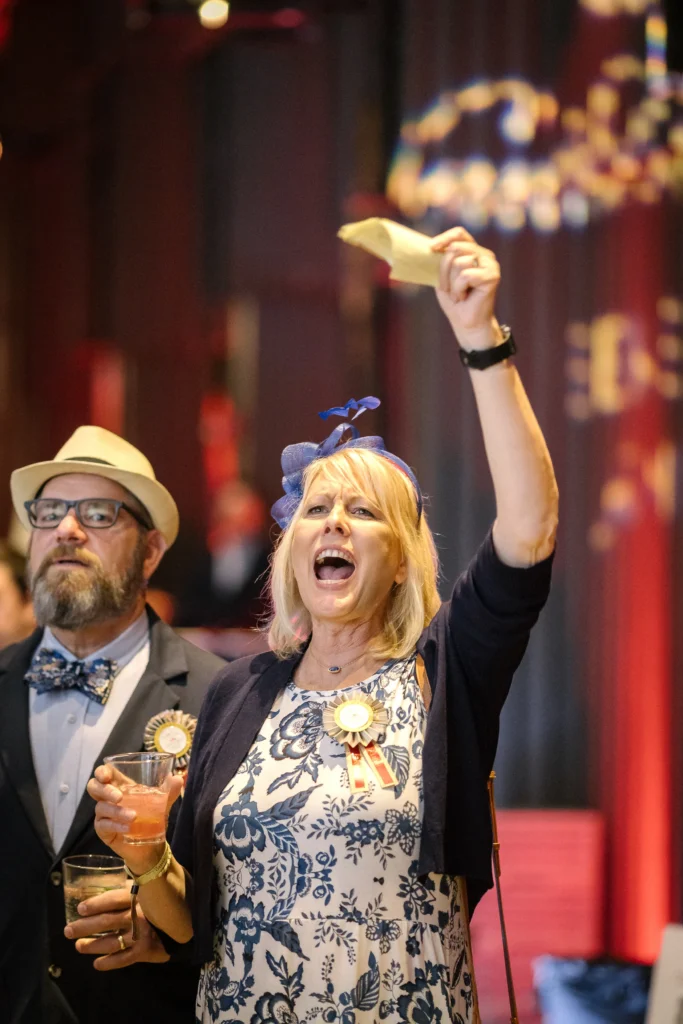 Woman in a blue floral dress cheers, raising a napkin high while holding a drink at a celebration (with a man nearby).  MitoAction