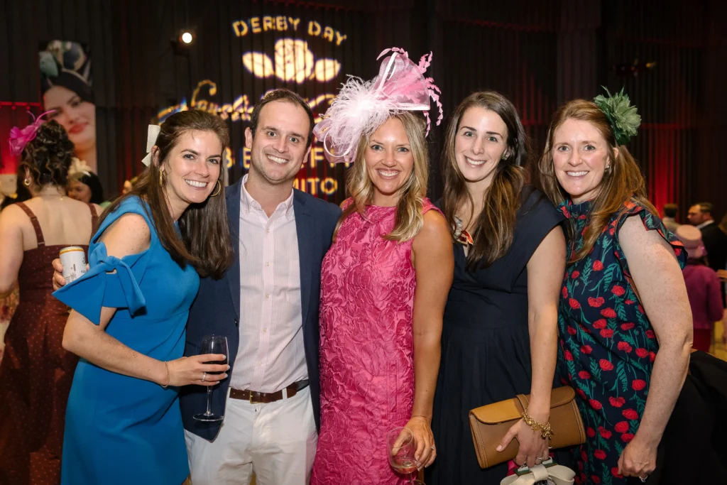 Five adults posing for a group photo at a Derby Day party, smiling and holding drinks, dressed in bright dresses and suits with hats/fascinators in the background.  MitoAction