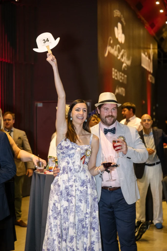 Woman in a floral dress raises a white hat marked 34 on a stick at a party, with a man in a light suit beside her holding a drink.  MitoAction