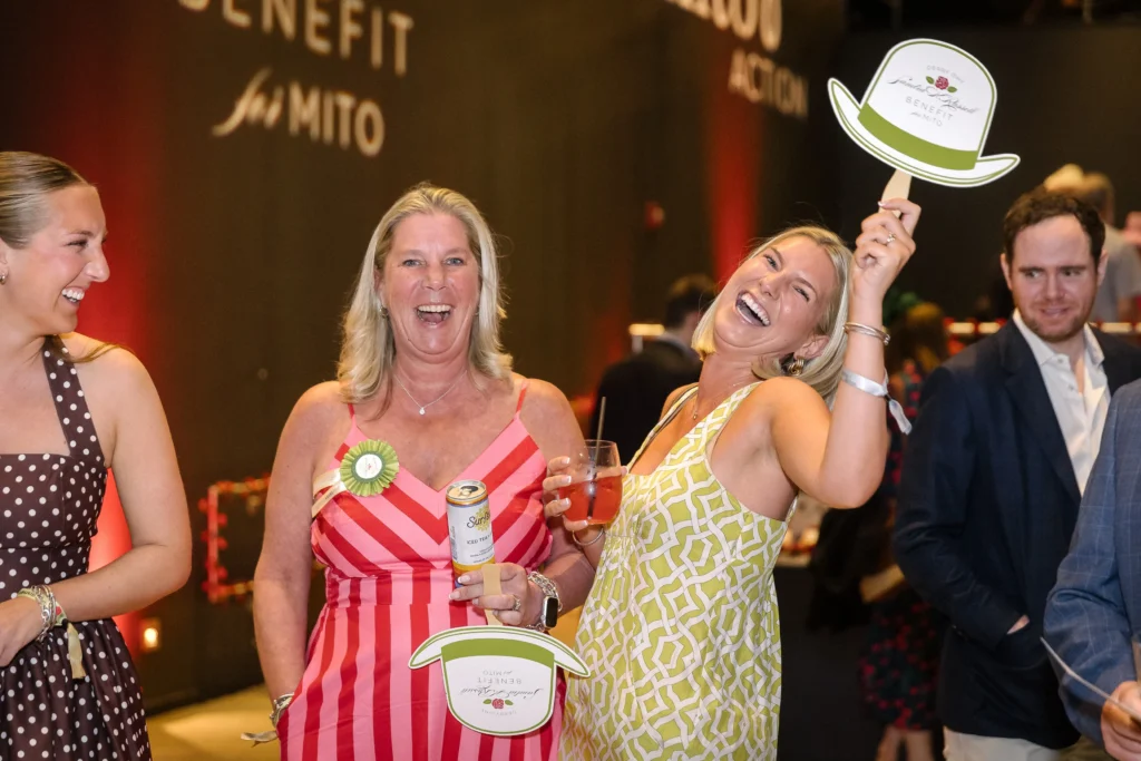 Group of friends celebrating at a party, smiling and toasting with drinks; woman in red-and-white striped dress holds a can and badge visible.  MitoAction
