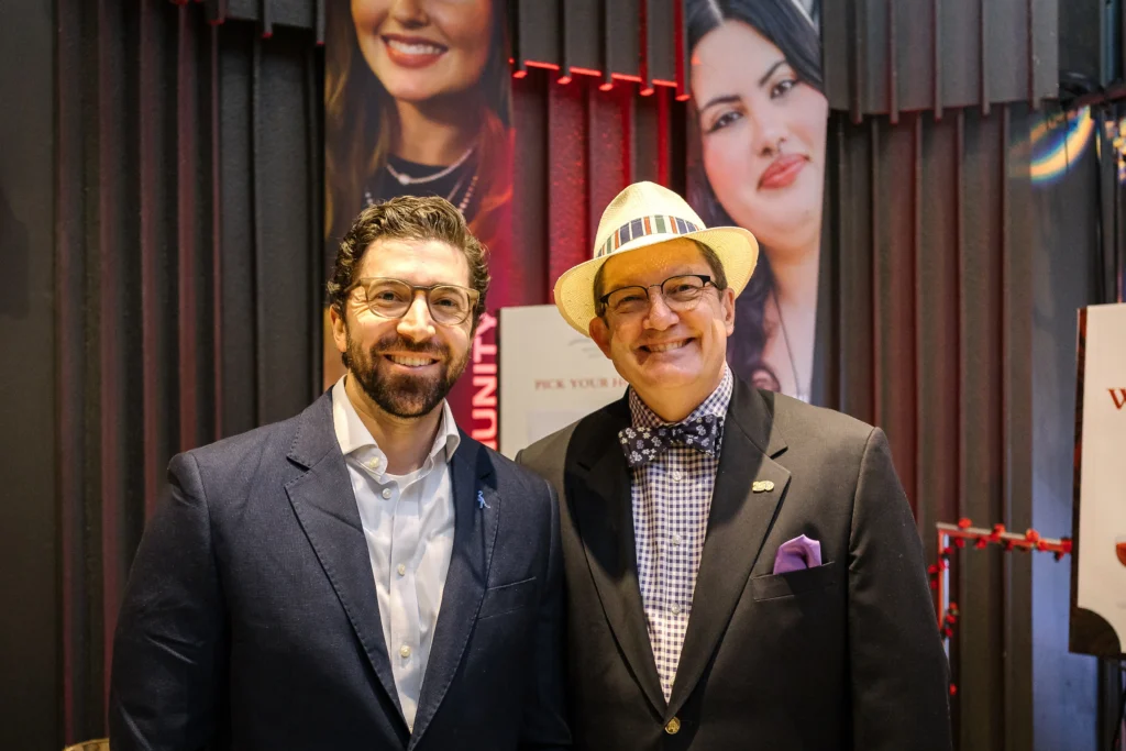 Two men in suits posing for a photo at an event, smiling, with large portrait banners behind them on dark curtains.  MitoAction