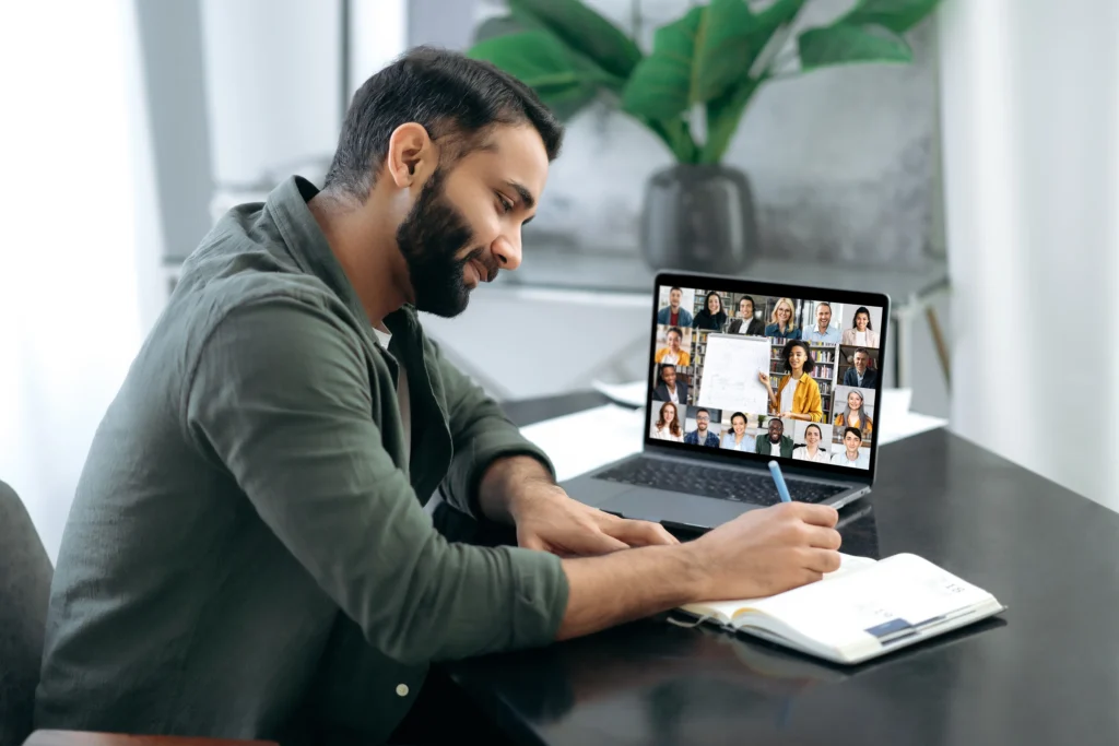 MitoAction Educational Series for Genetic Counselors A Man Taking Notes While on a Virtual Webinar - MitoAction Man sits at a desk taking notes while participating in a video conference on a laptop showing multiple participants in a grid view. MitoAction